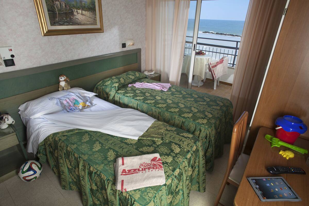 A hotel room with two beds, a balcony with a table and chairs, a small table with a remote control, and a stuffed toy on the left side.