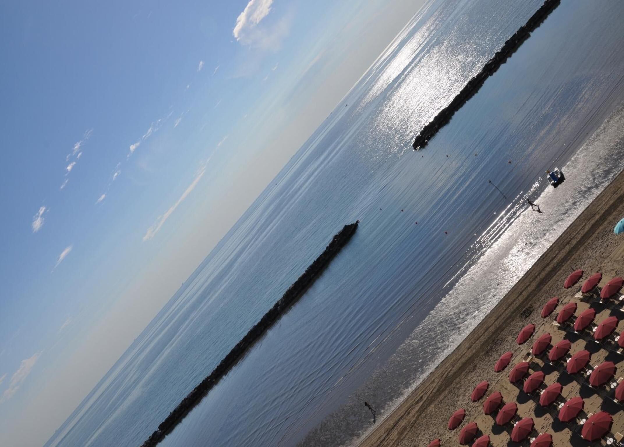Ein Strand mit blauem Himmel, ruhigem Wasser und einer Reihe roter Sonnenschirme im Sand.