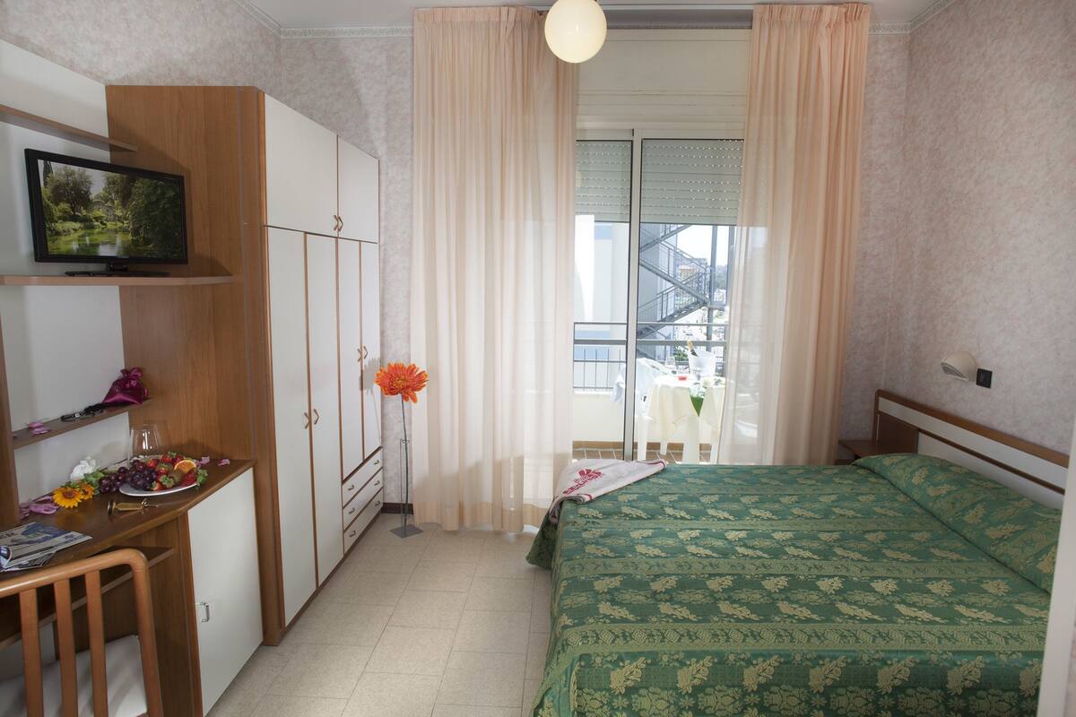A hotel room with a bed, a desk with a computer, a TV, and a window with curtains.