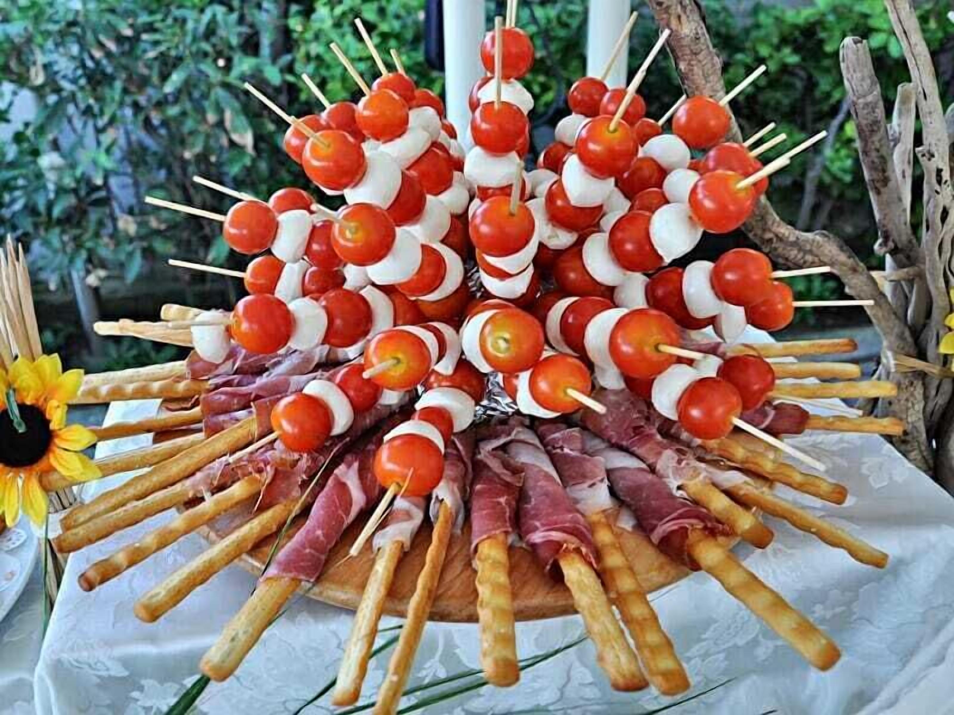Esposizione di cibi con spiedini di pomodori e prosciutto, disposti su una tovaglia bianca.