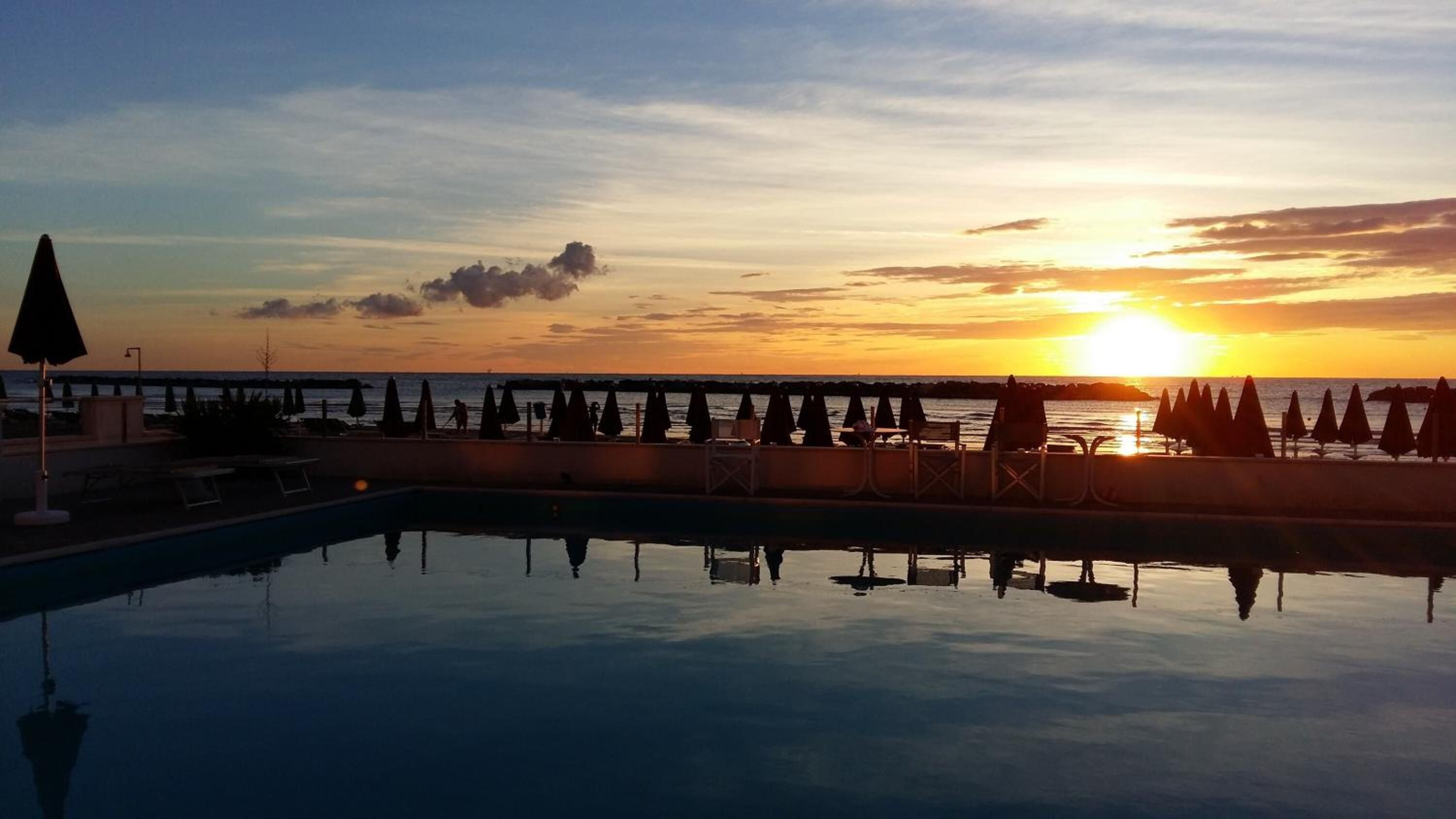 Une piscine avec vue sur le coucher de soleil, des chaises longues et des parasols disposés le long du bord de la piscine.