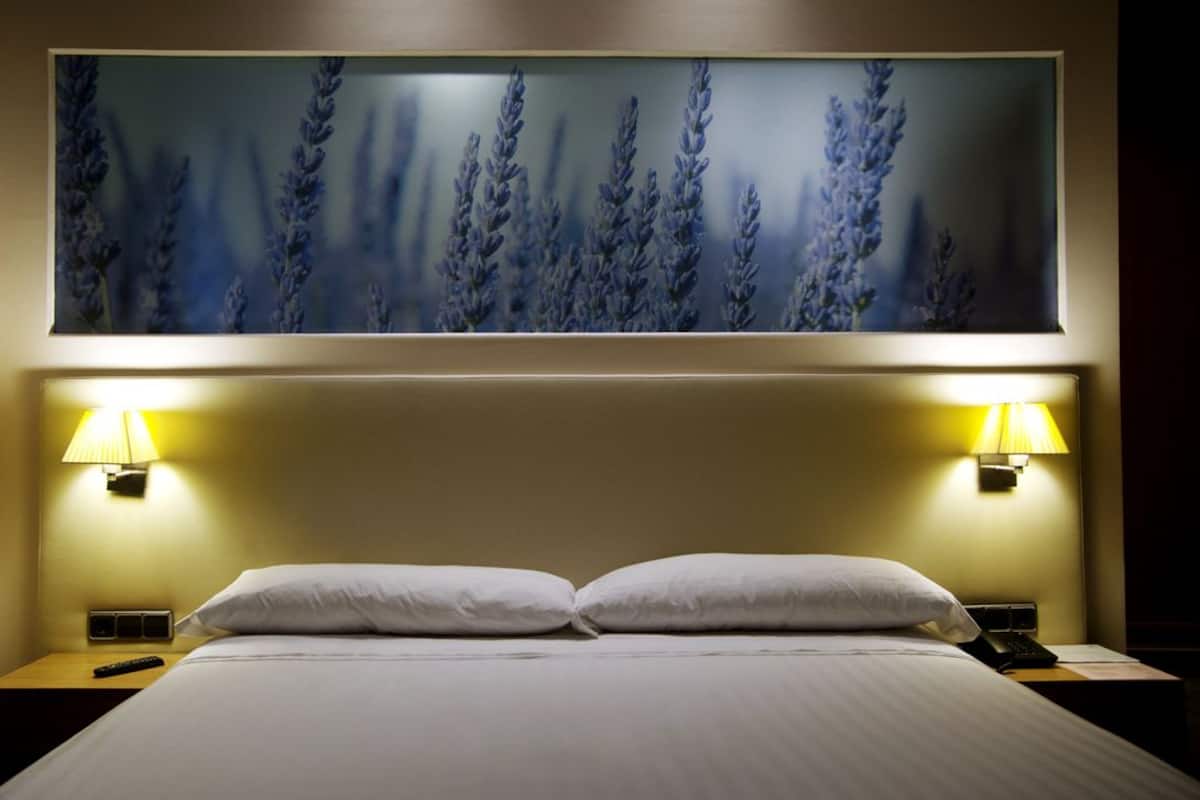 Quarto de hotel com uma cama grande, duas luminárias de cabeceira, uma mesa de apoio e um quadro de lavanda fixado na parede.