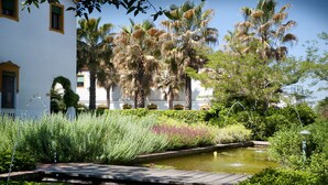 Un jardín con un estanque, abundante follaje y un edificio blanco con detalles amarillos.