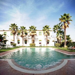 A pool area with a clear blue pool, surrounded by palm trees and white buildings with green windows.