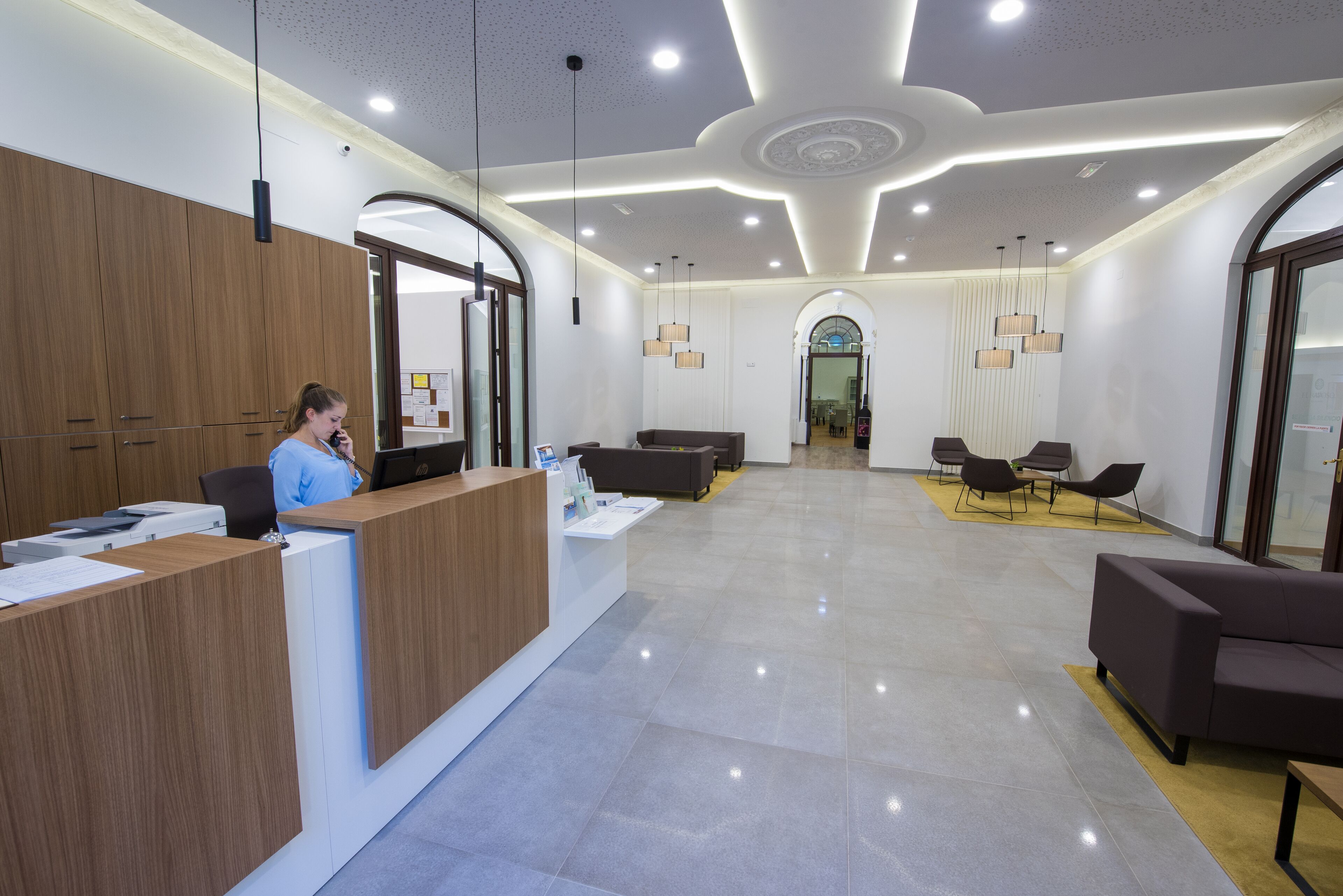Un área de recepción moderna con un mostrador de madera, una zona de estar con sofás y sillones, y un espejo grande sobre el mostrador.