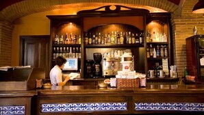 Una zona de bar con estantes llenos de botellas, una máquina de café y una barra donde trabaja una persona.