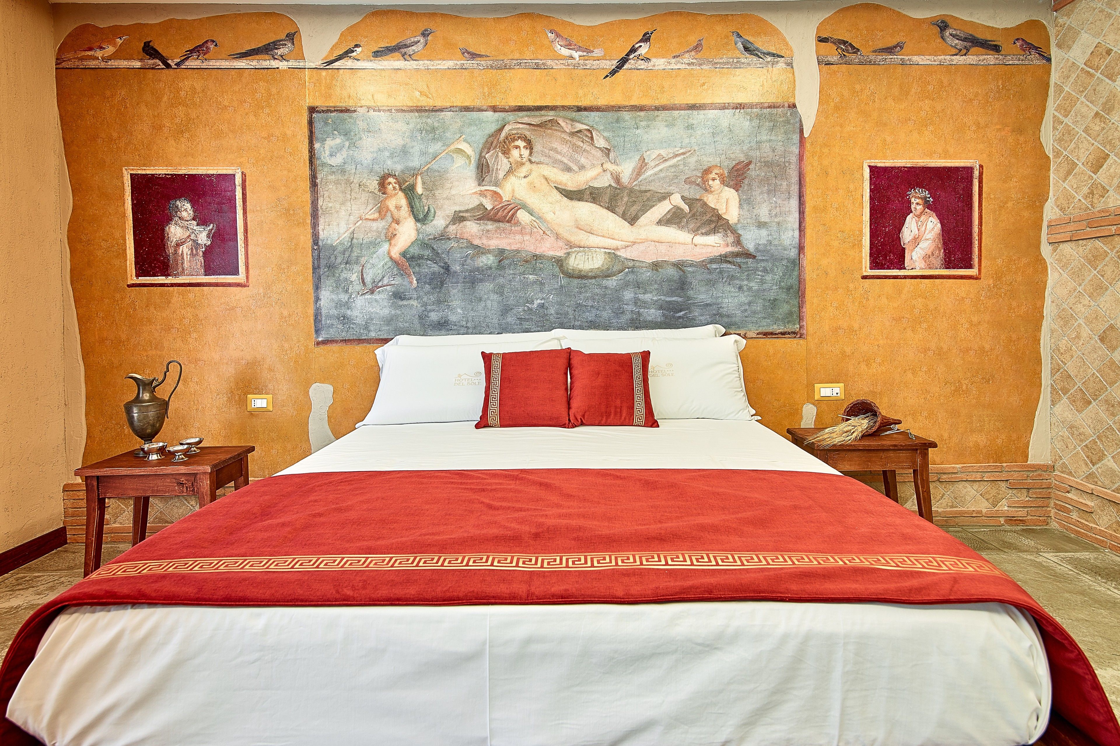 Une chambre d’hôtel avec un grand lit, une literie rouge et blanche, une fresque murale et deux tables de chevet avec des lampes.