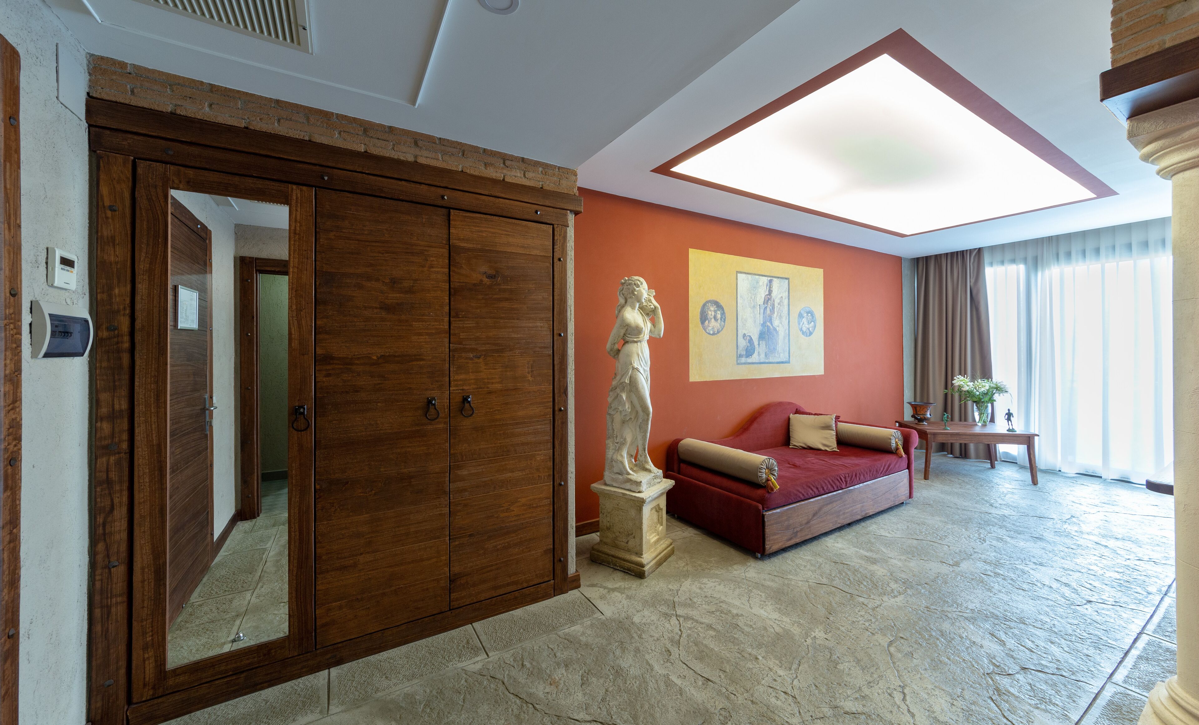 Um quarto espaçoso com um guarda-roupa de madeira grande, um sofá vermelho, uma estátua e um claraboia.
