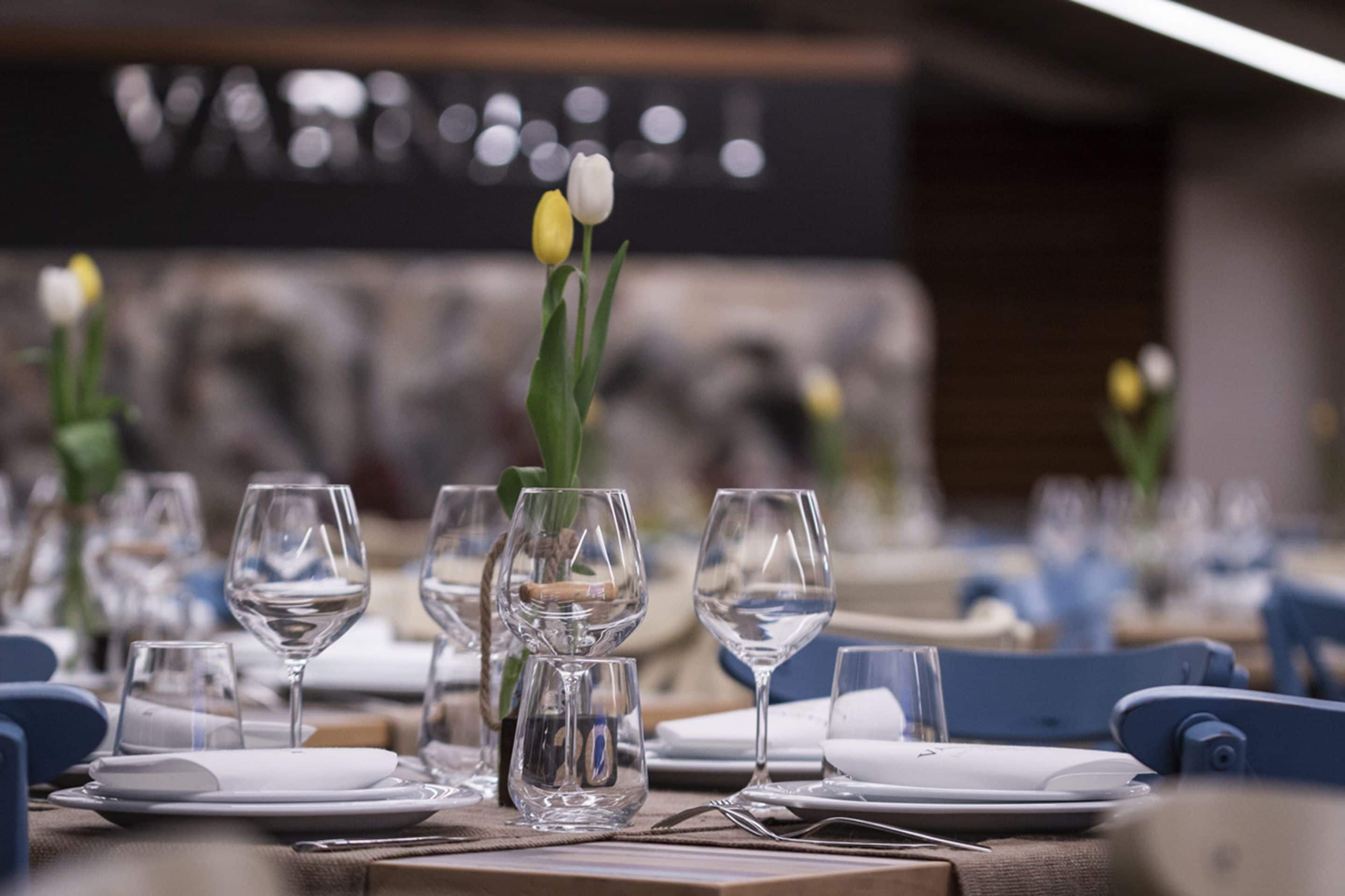 A dining table set with clear glassware, plates, and cutlery, with a vase of tulips in the center.