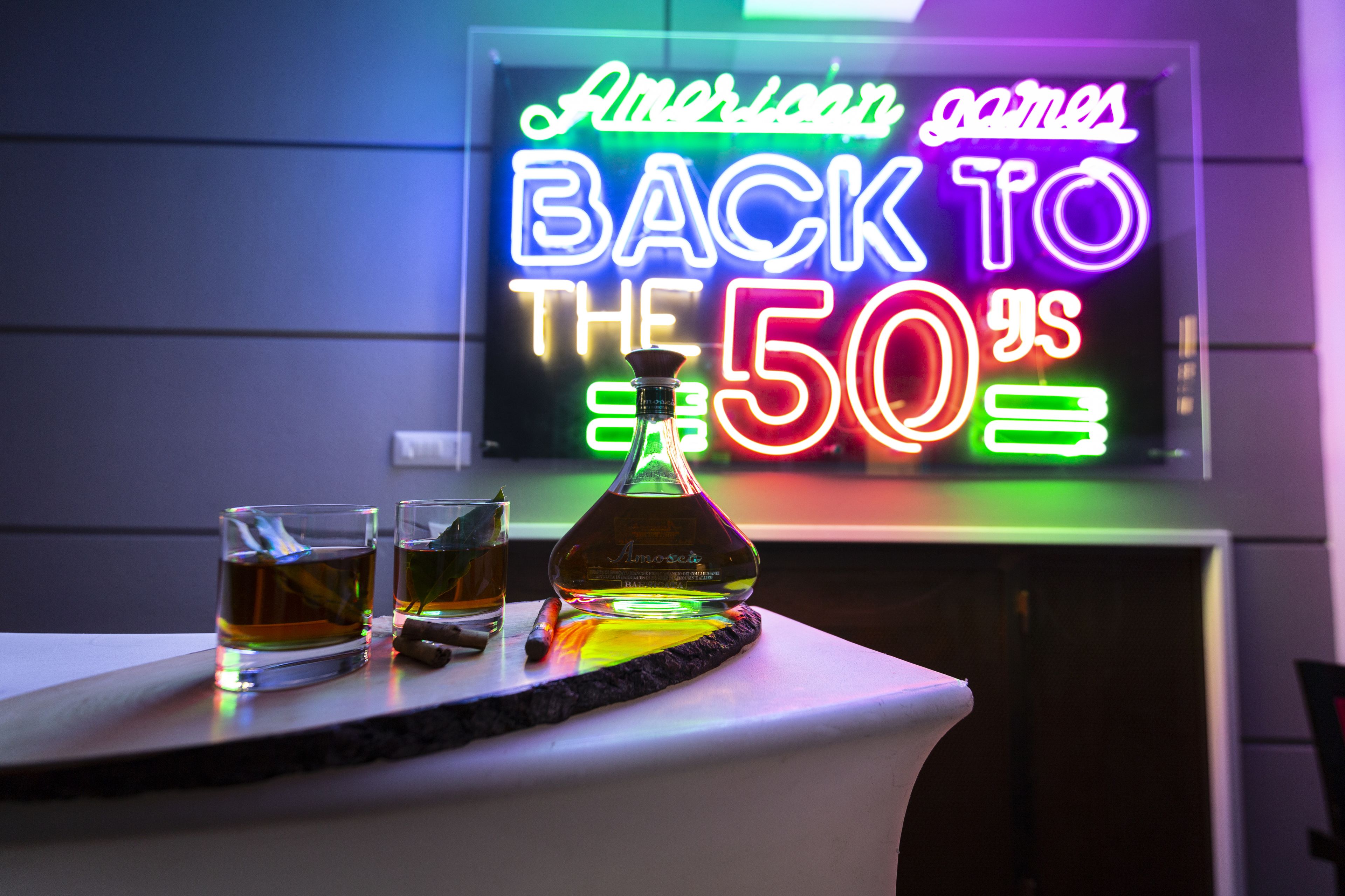 A bar setting with a neon sign, two glasses of whiskey, and a bottle of Amosite whiskey on a table.