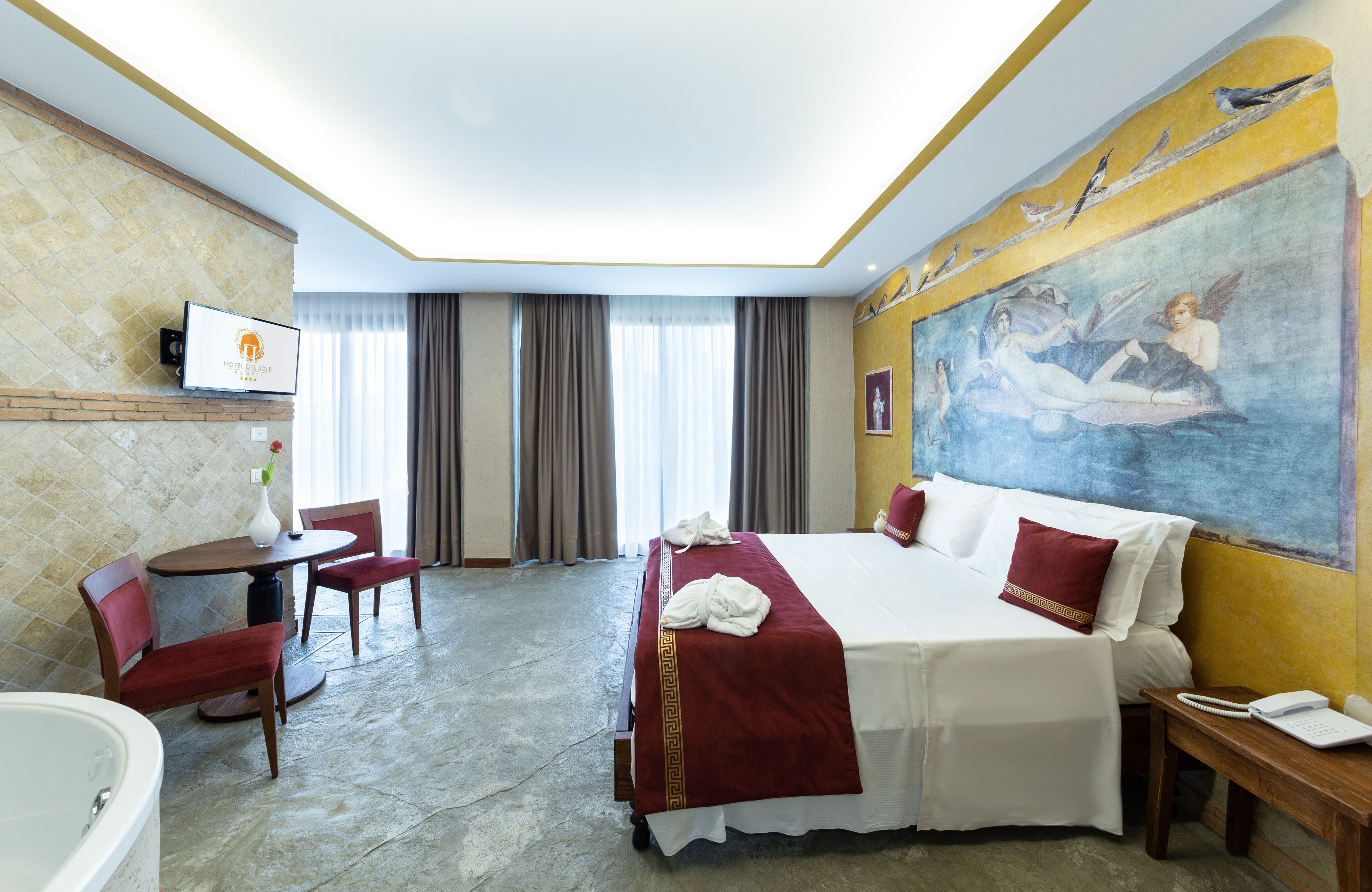A hotel room with a large bed, a desk with a chair, a small table, a TV mounted on the wall, and a painting on the wall.