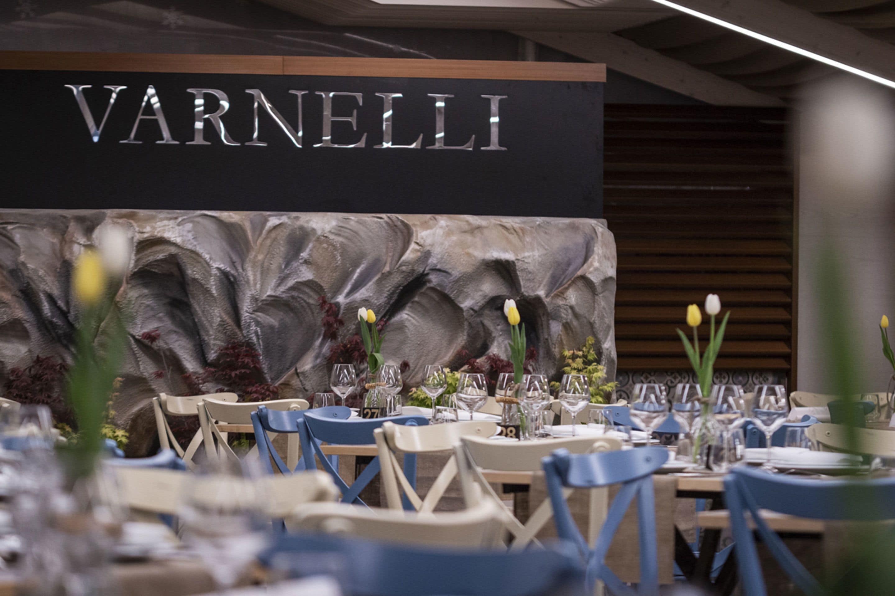 A restaurant interior with a black wall displaying 'VARNELLI' in metallic letters, tables set with chairs, and floral centerpieces.