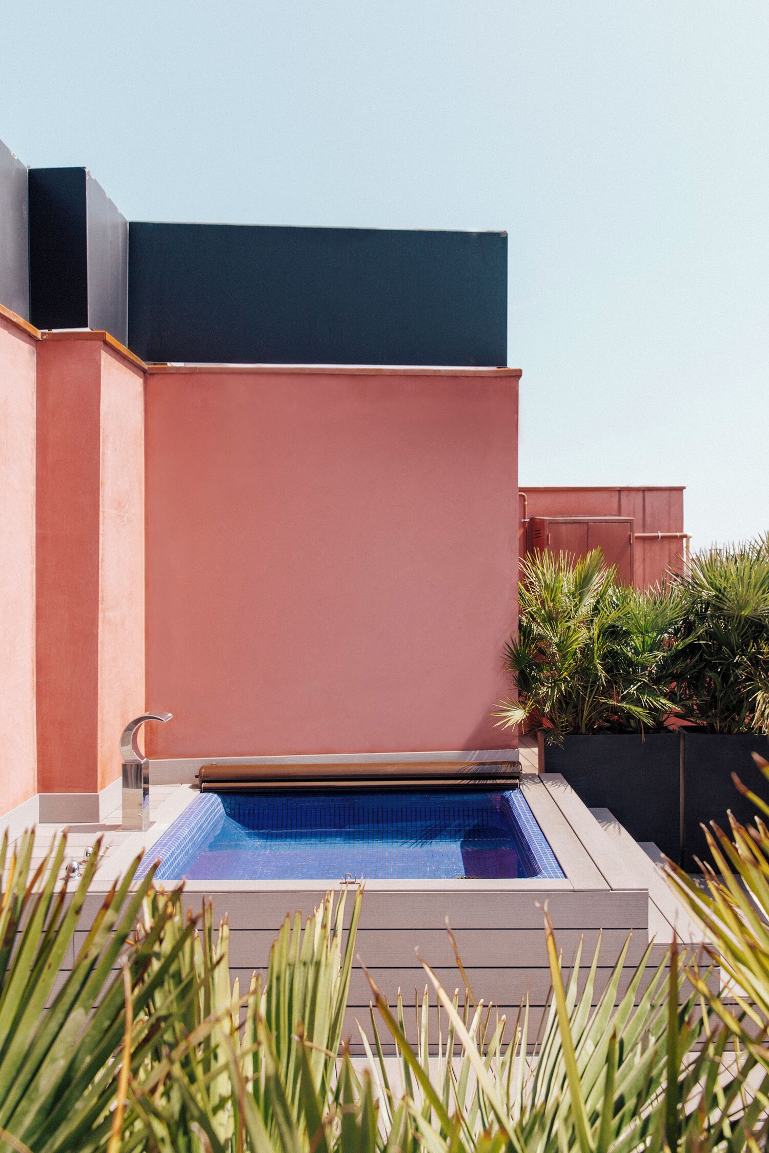 A swimming pool with a tiled deck, surrounded by a building with a flat roof and a pink wall.