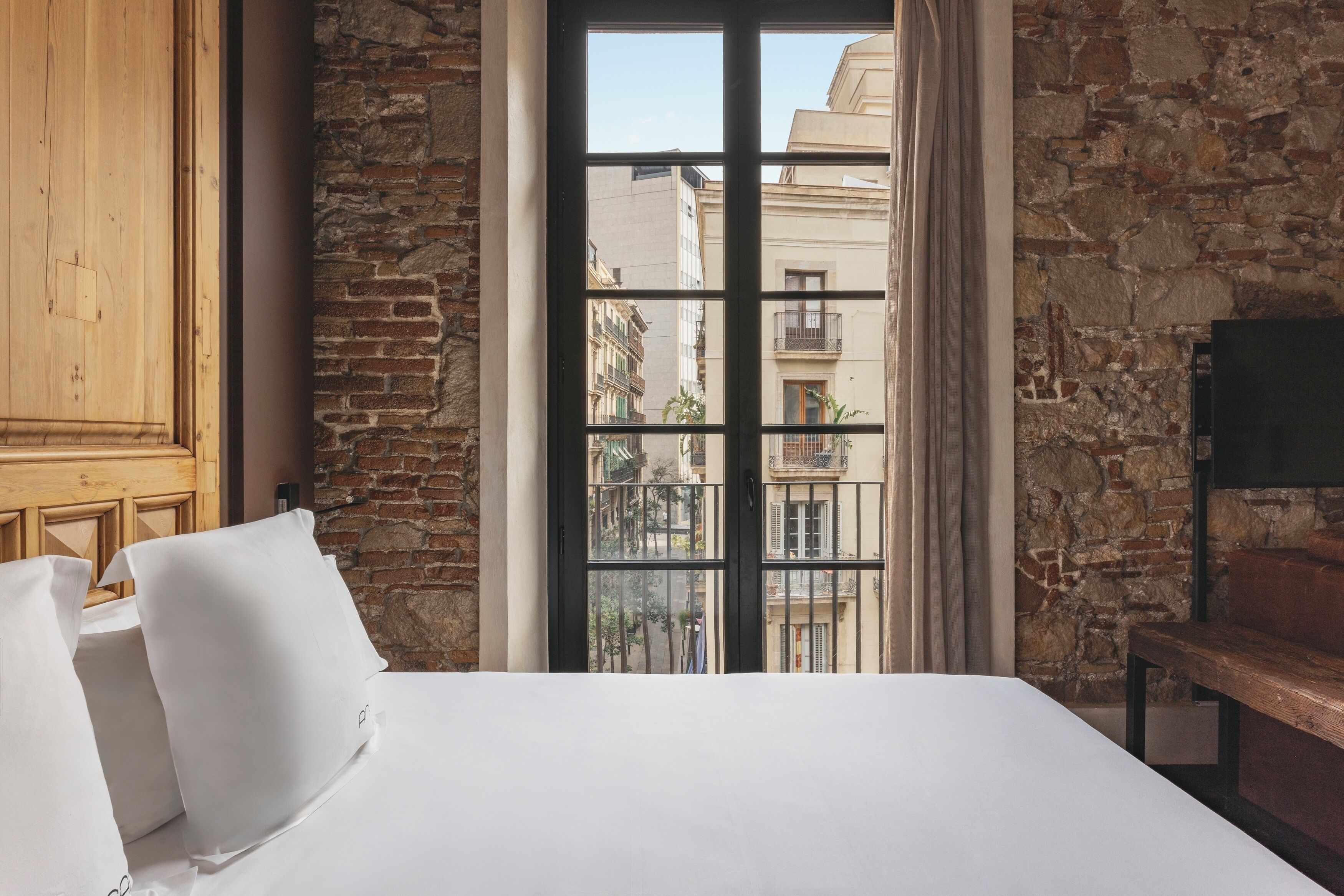 Habitación de hotel con una cama grande, un armario de madera, una pared de piedra y vistas a edificios a través de una ventana.