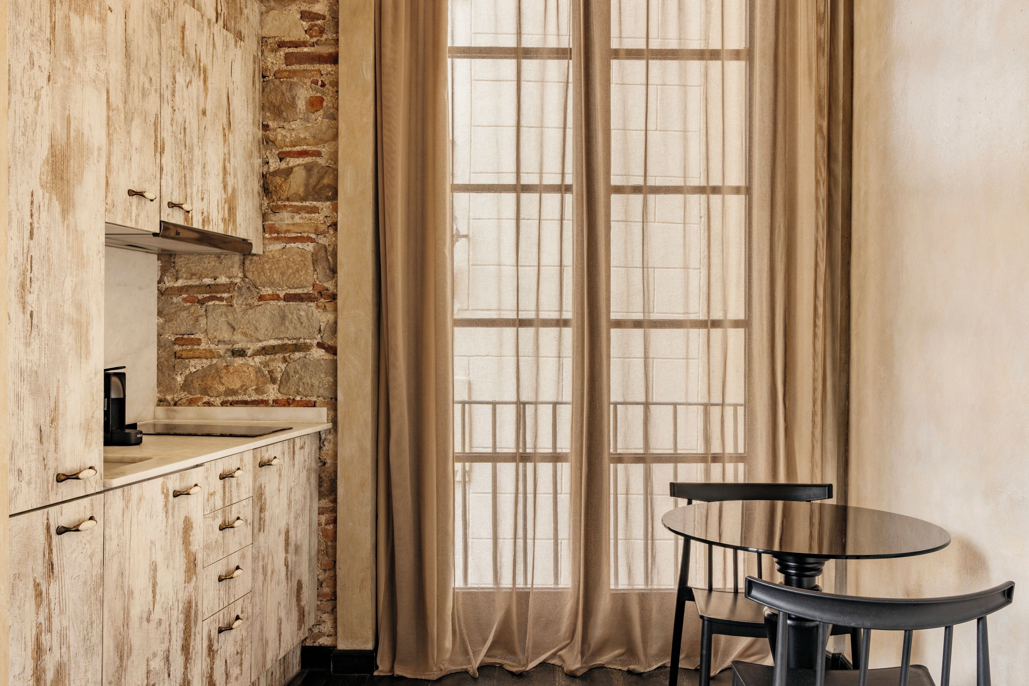 Une cuisine avec un mur en pierre, une fenêtre avec des rideaux et une petite table ronde entourée de chaises.