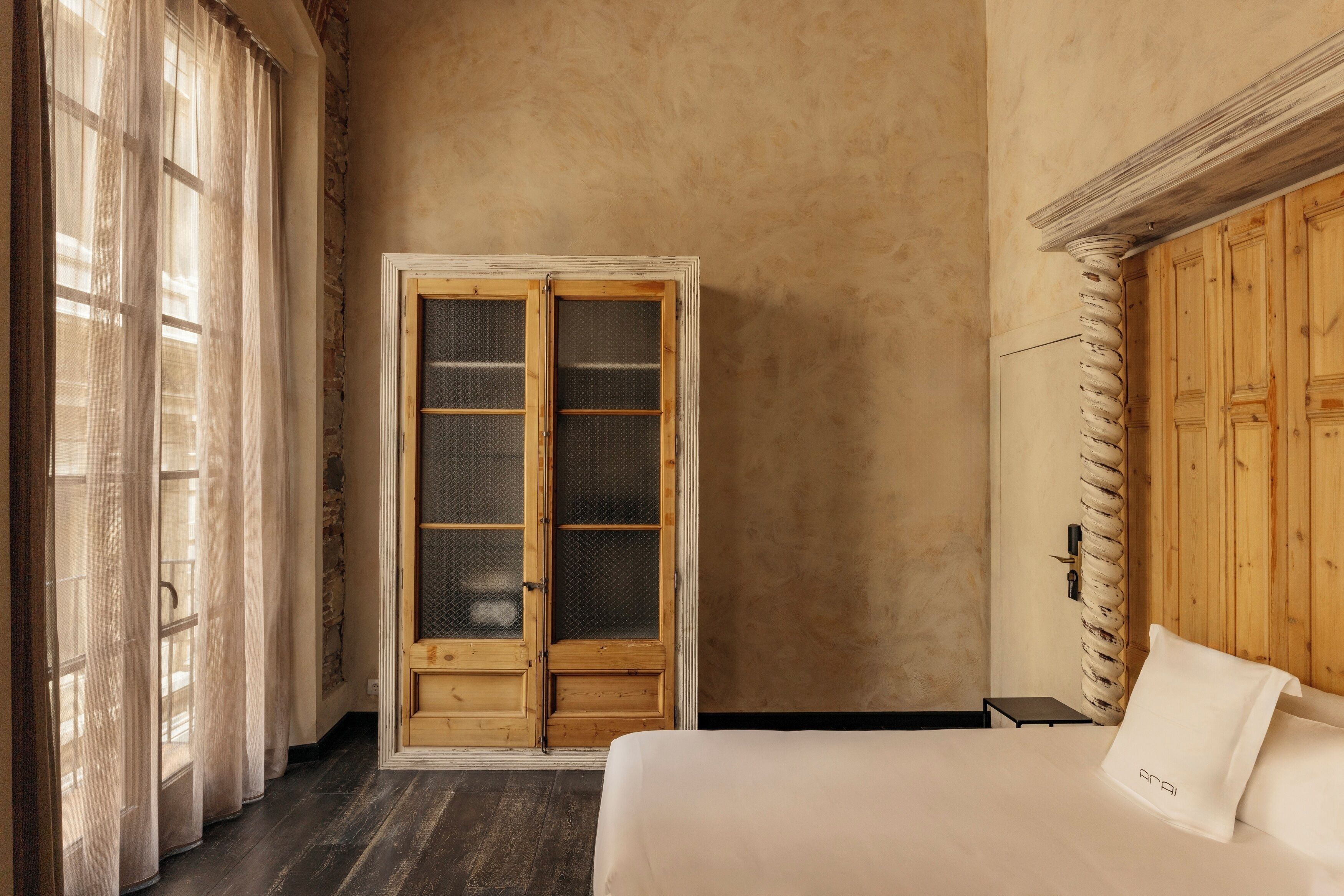 A room with a bed, a wooden wardrobe, a window with curtains, and a wooden floor.