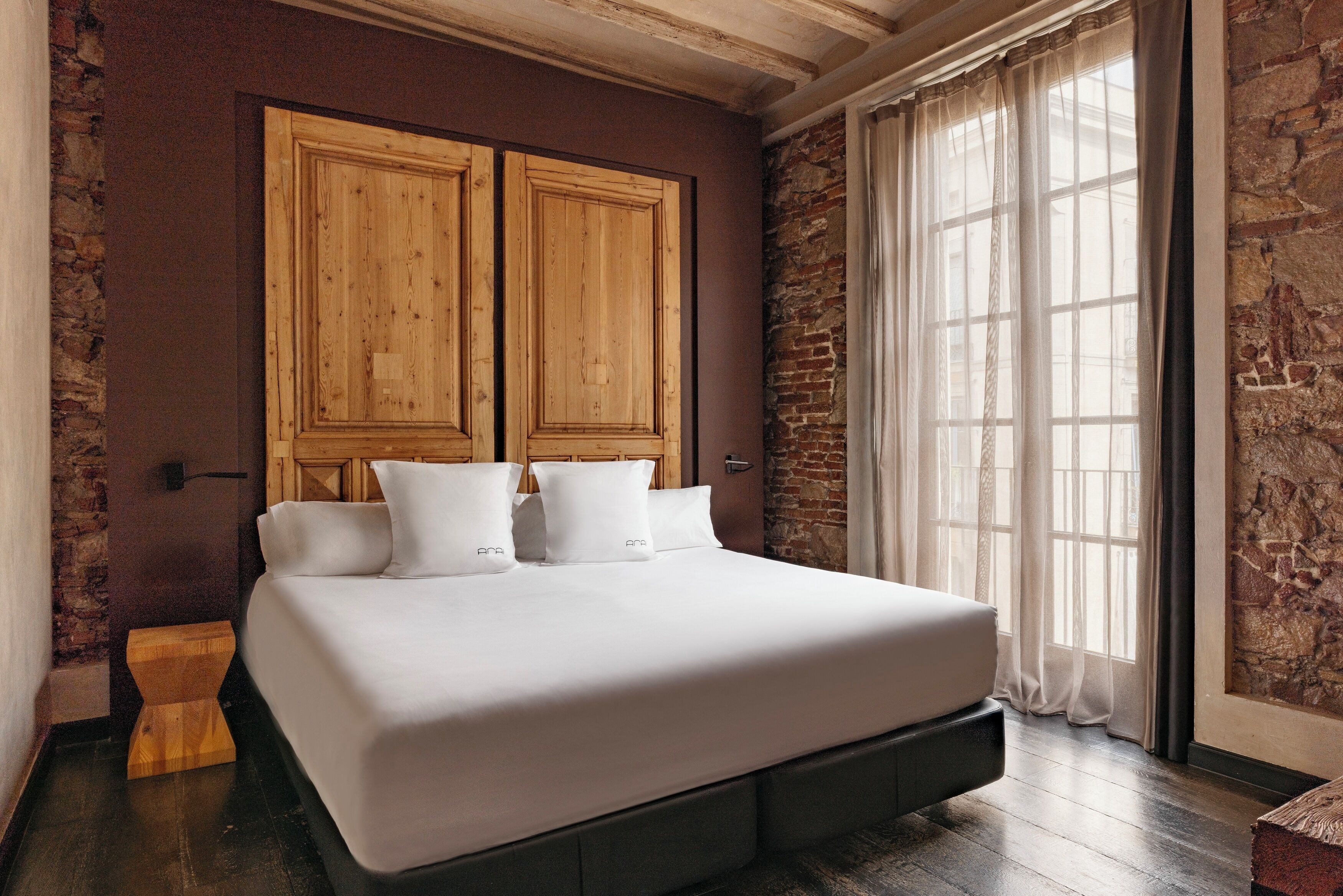 A bed with a wooden headboard, a stone wall, and a large window with curtains.