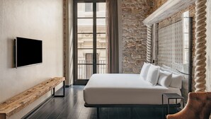 A bedroom with a bed, a bench, a TV, and a window with a view of buildings. - Arai Aparthotel (Barcelona)