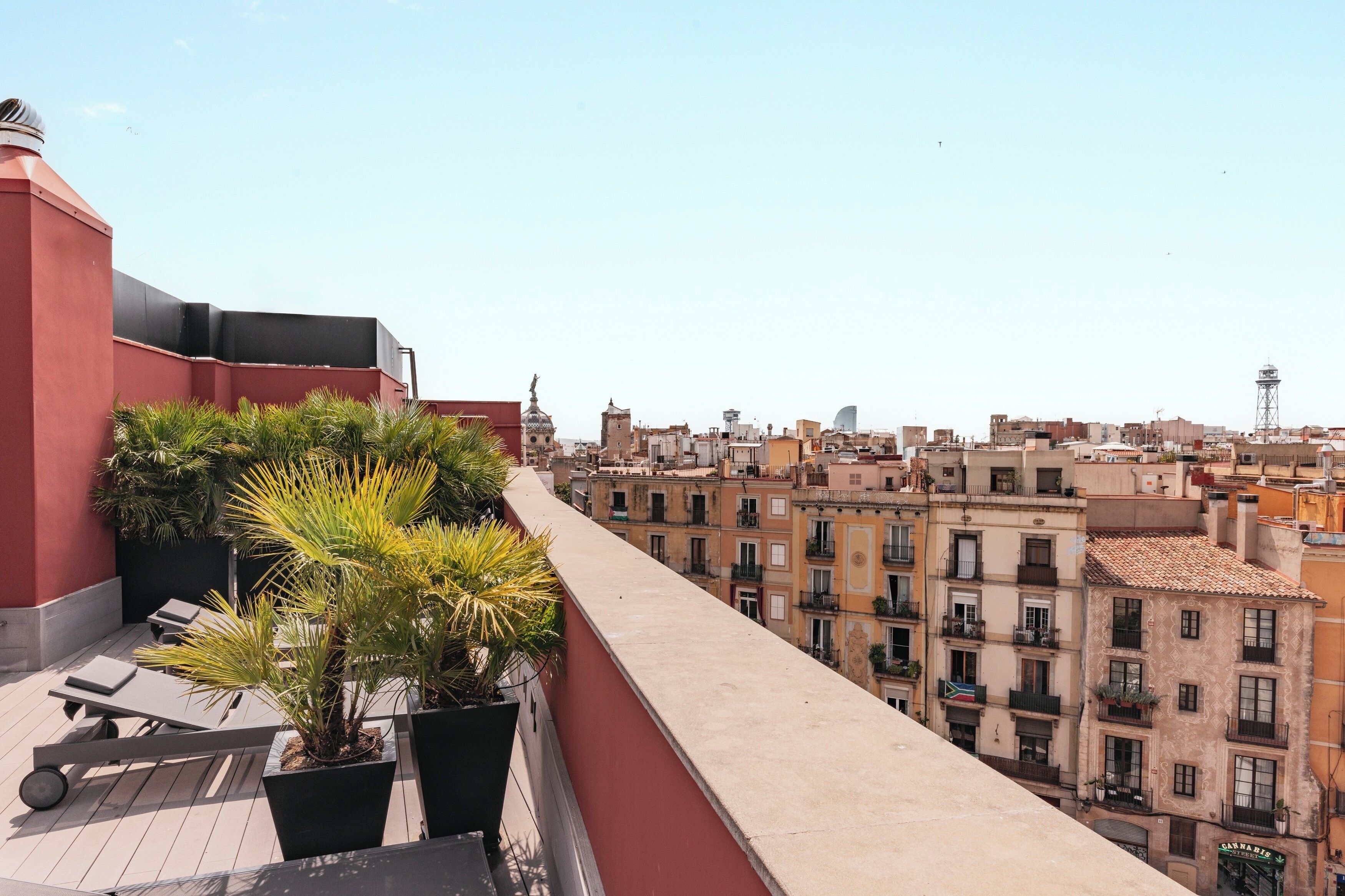 Terraço no topo do prédio com plantas em vasos, uma espreguiçadeira e vista para um horizonte urbano com edifícios e uma torre.