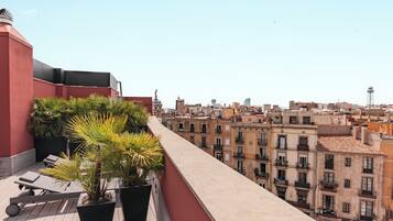 Terraço no topo do prédio com plantas em vasos, uma espreguiçadeira e vista para um horizonte urbano com edifícios e uma torre.