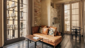 A room with a leather sofa, a small table, and a view of a building through large windows. - Arai Aparthotel (Barcelona)