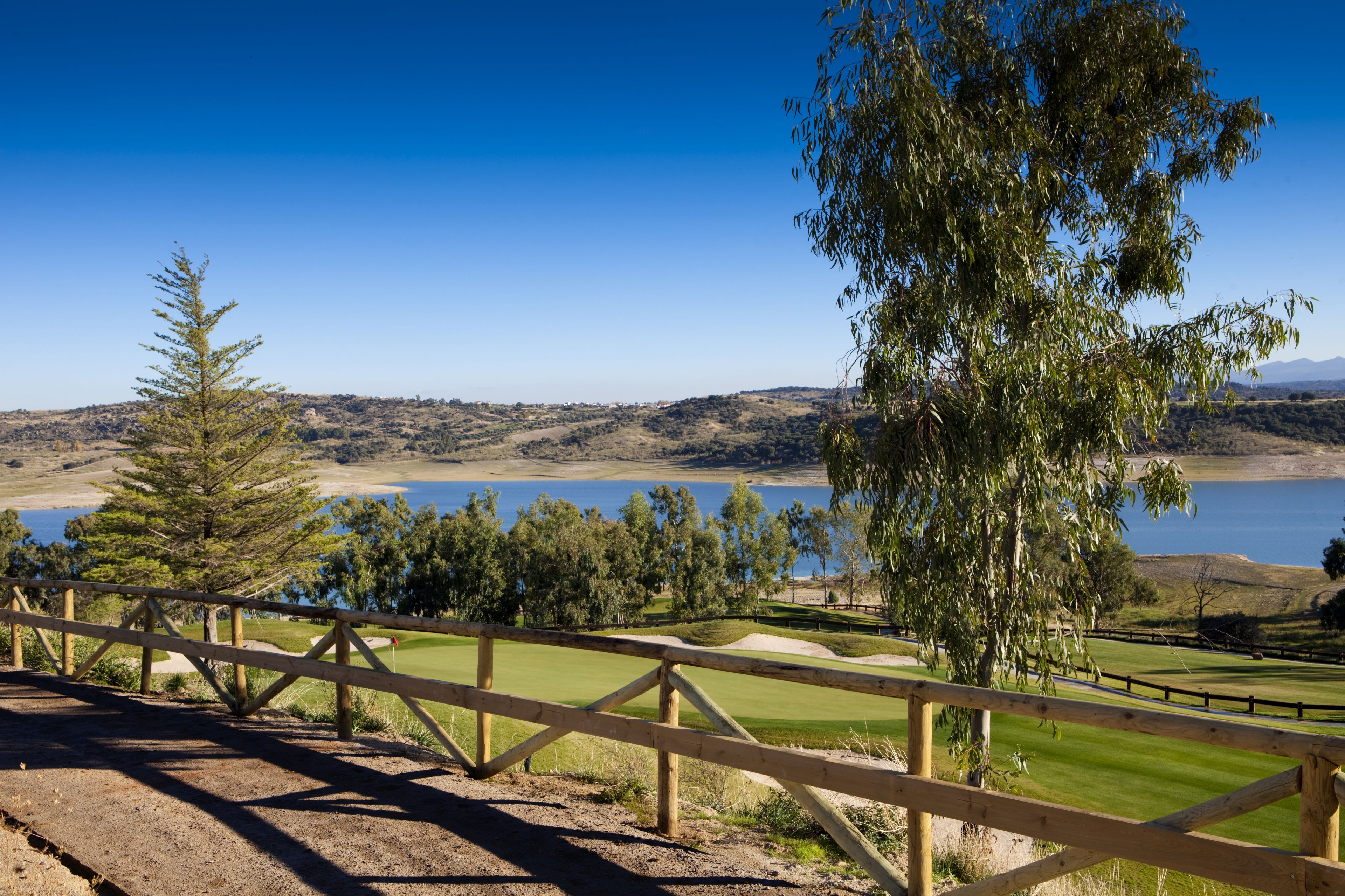 A golf course with a wooden fence, trees, and a view of a lake and hills in the background.
