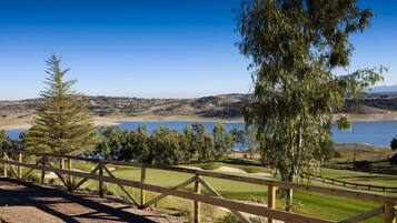 Campo de golf con valla de madera, árboles y vistas a un lago y colinas al fondo.