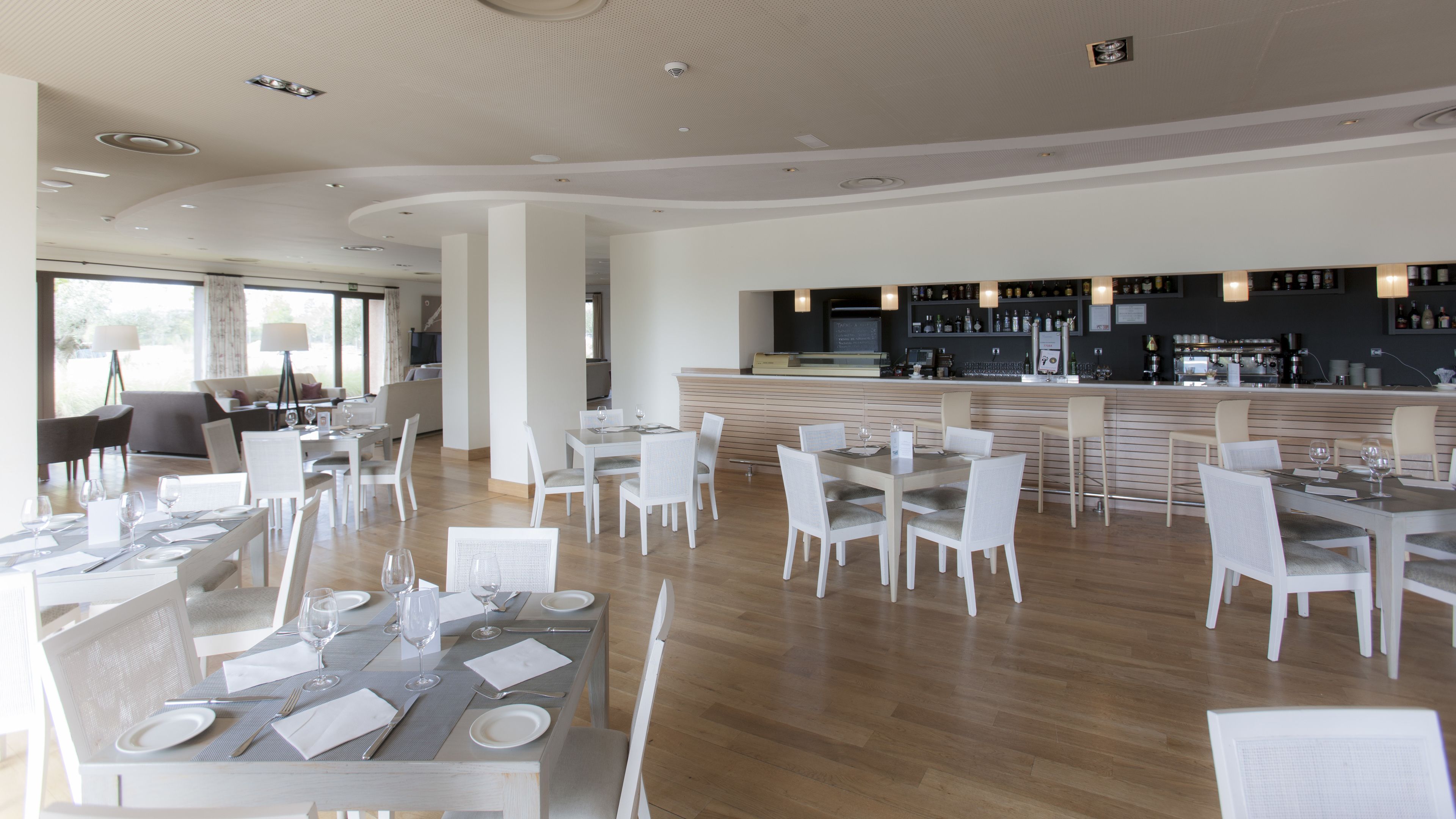 A modern restaurant with white furniture, a bar area, and a view of the outdoors.