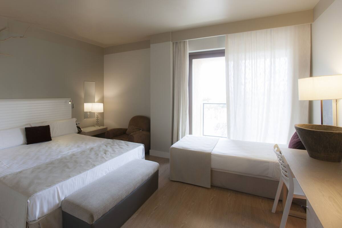A hotel room with a large bed, a smaller bed, a desk, a chair, a nightstand, a lamp, a mirror, and a window with curtains.