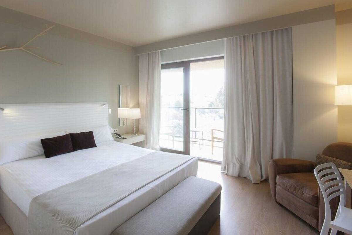 A hotel room with a large bed, a desk with a lamp, a chair, and a view of the outdoors.