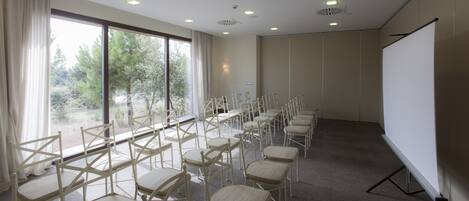 Un salón de conferencias con filas de sillas, una pantalla de proyector y grandes ventanales con vistas a árboles.