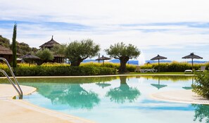 Una piscina con sillas de descanso y sombrillas, rodeada de vegetación y chozas tradicionales.
