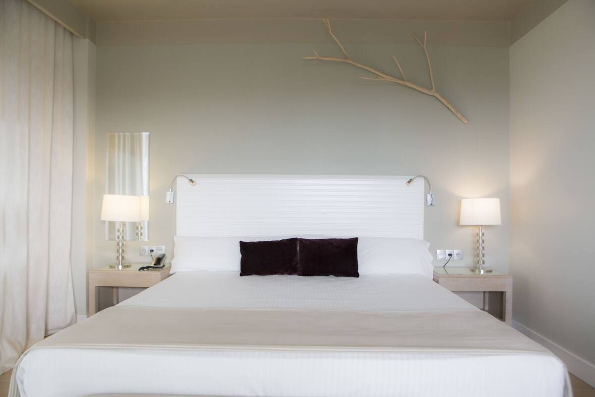 A hotel room with a large bed, two bedside tables with lamps, a wall-mounted branch decoration, and a window with curtains.