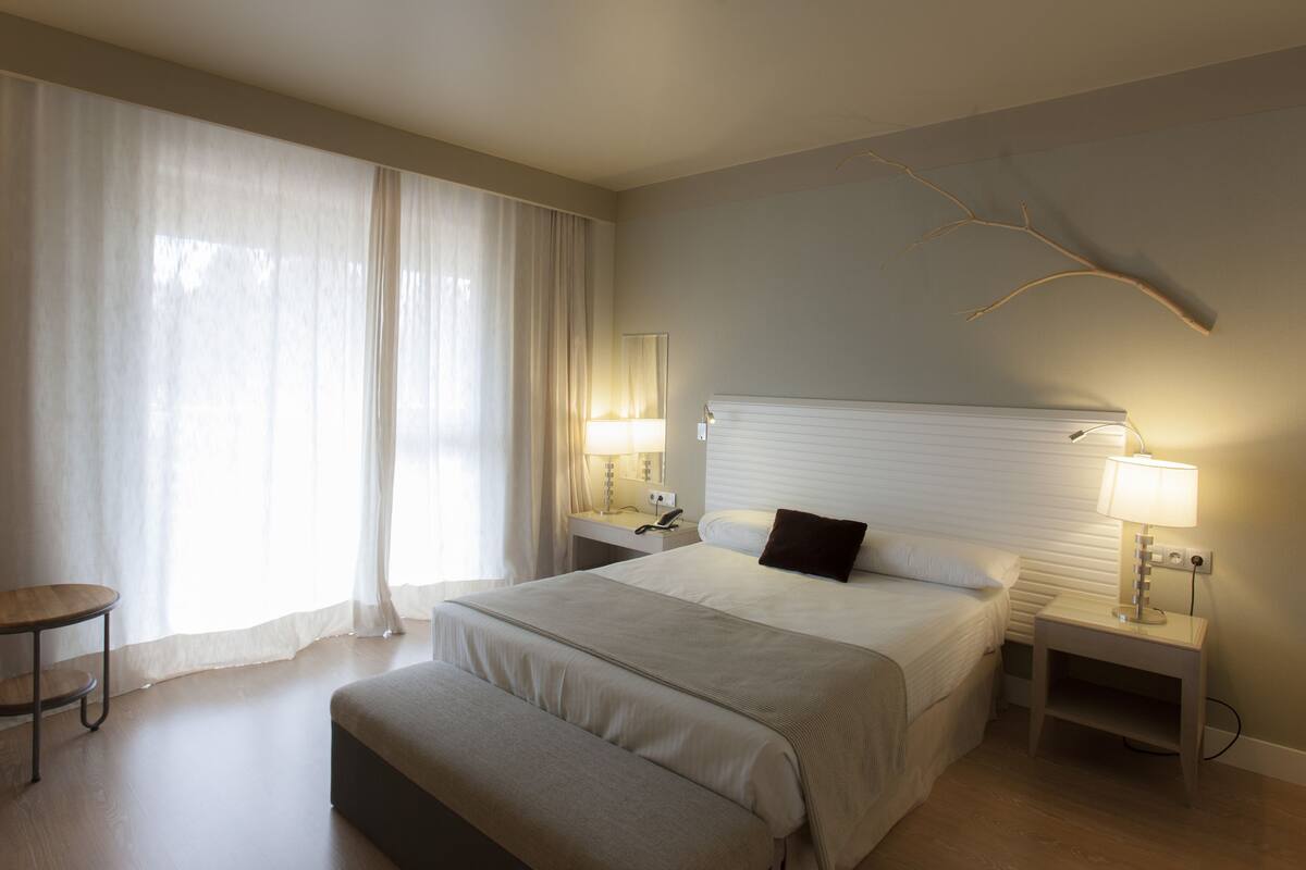 A hotel room with a large bed, a bedside table, a chair, and a window with curtains.