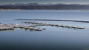 A marina with multiple docks extending into calm waters, surrounded by a hilly landscape.
