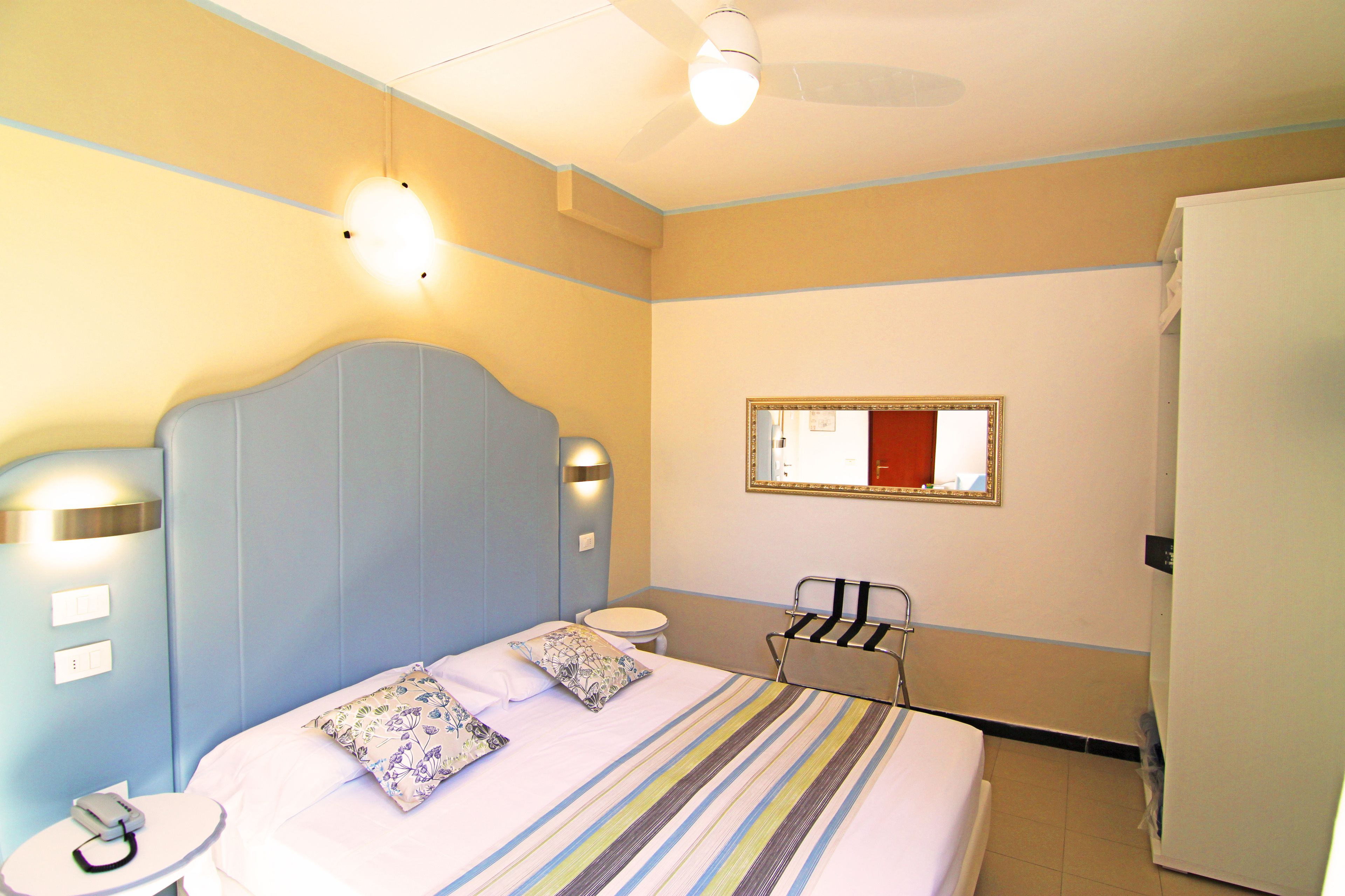 A hotel room with a bed, bedside table, lamp, mirror, and a refrigerator.