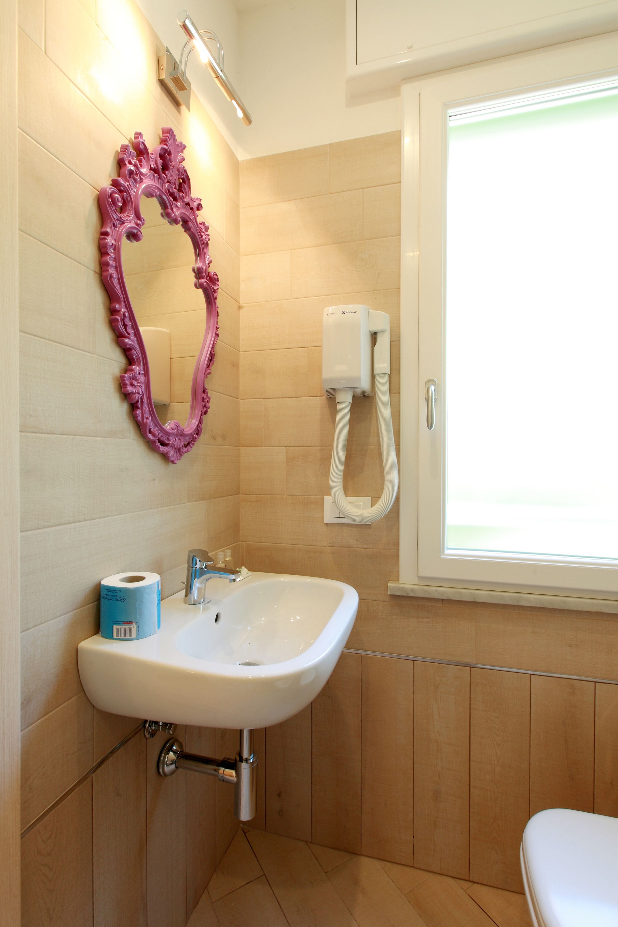 Une salle de bain avec un lavabo blanc, un miroir rose à motifs décoratifs et une fenêtre avec un distributeur de serviettes.