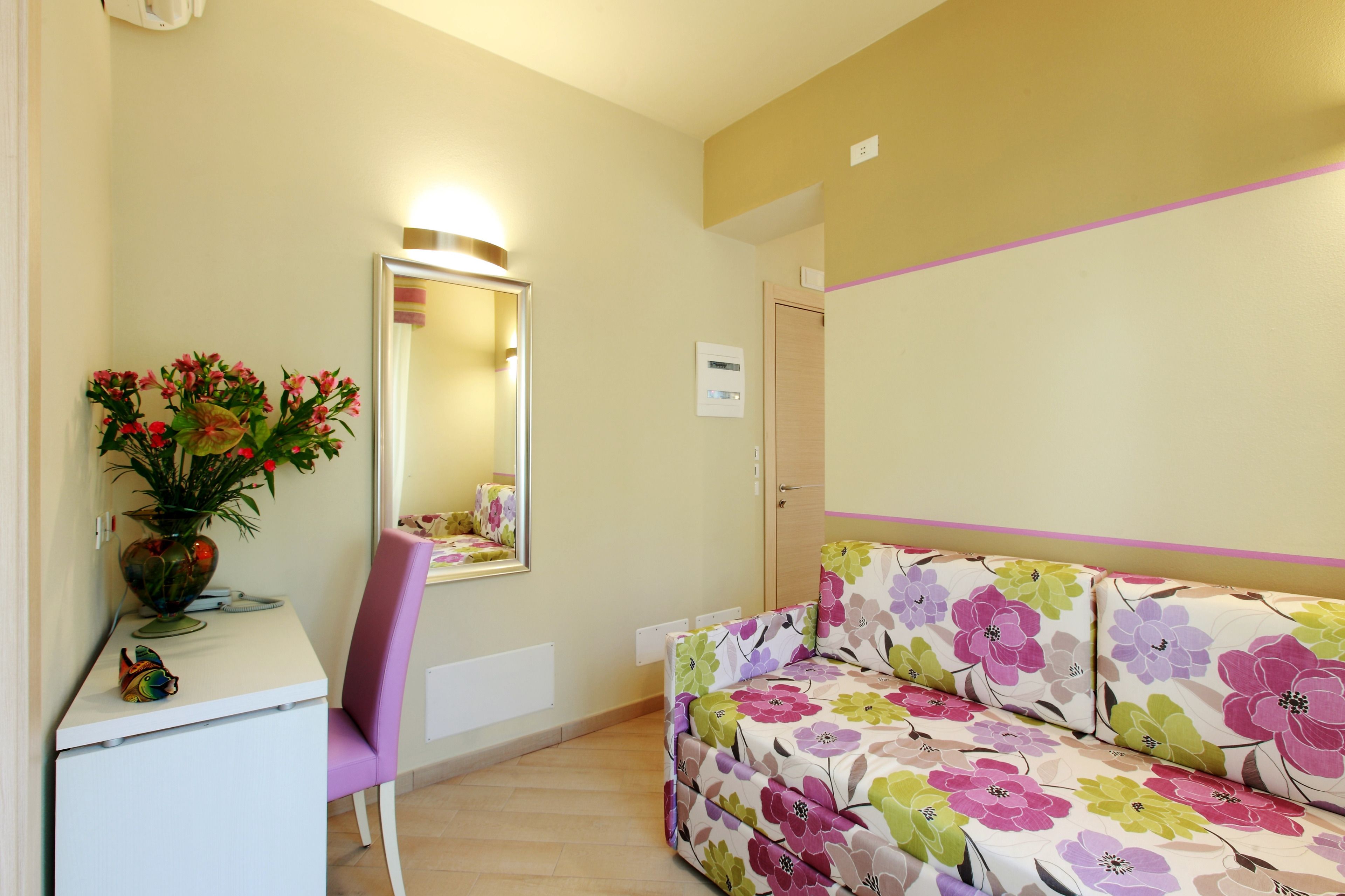 A room with a white desk, a chair, a floral sofa, a mirror, and a wall-mounted thermostat.