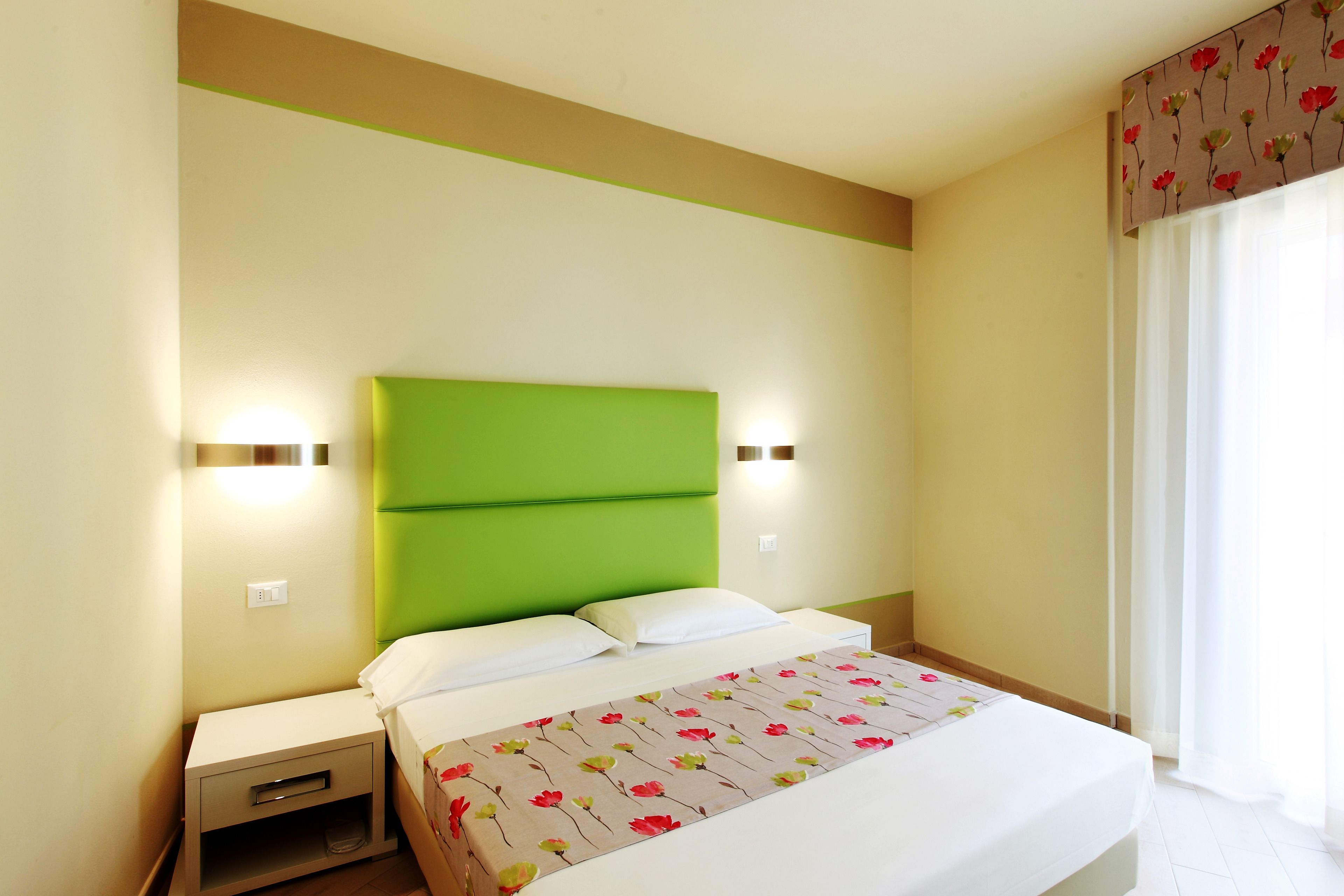 Una habitación de hotel con cabecera verde, ropa de cama blanca y una colcha estampada con motivos florales.