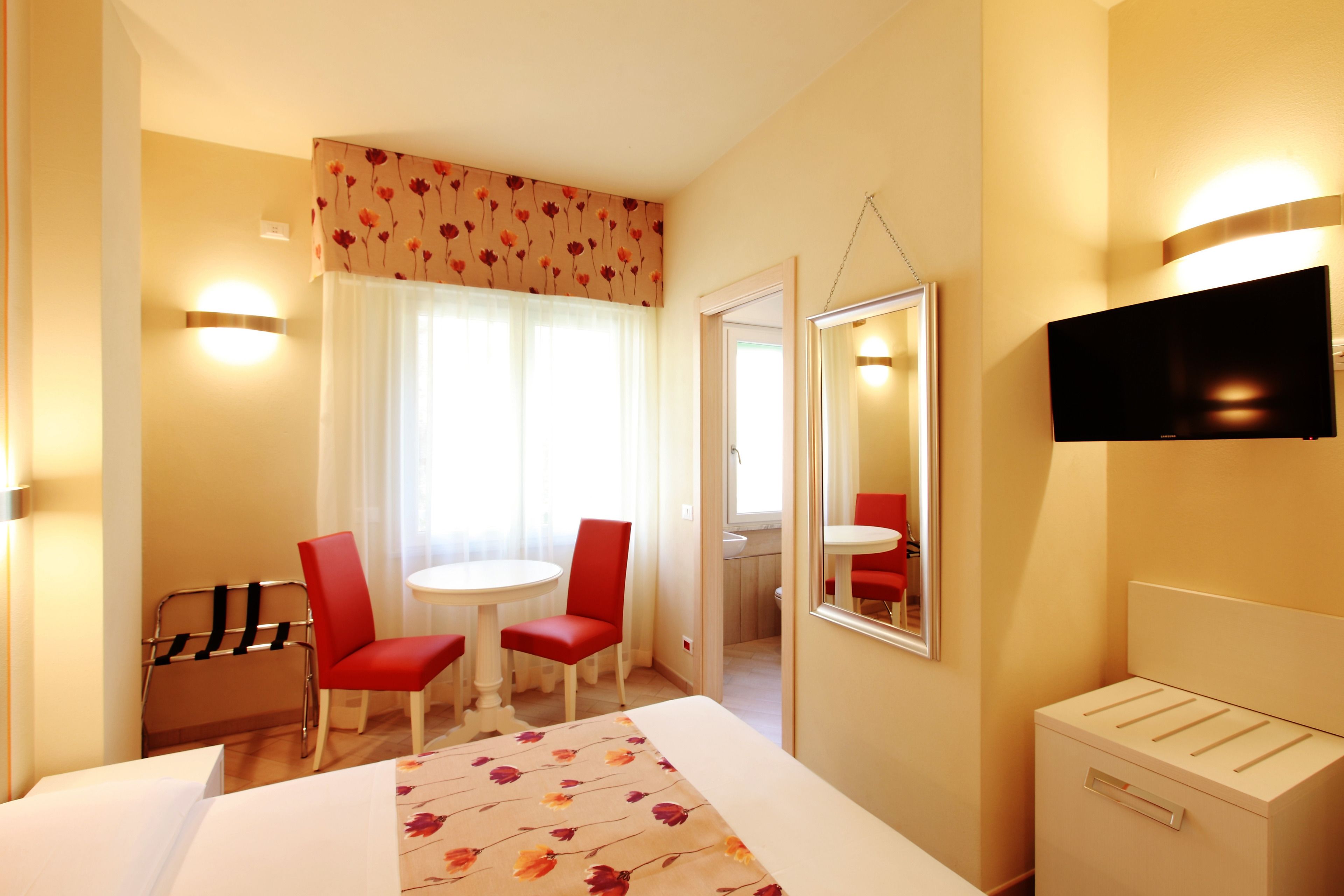 Una habitación de hotel con una cama, dos sillas rojas, una mesita, un televisor y un espejo.