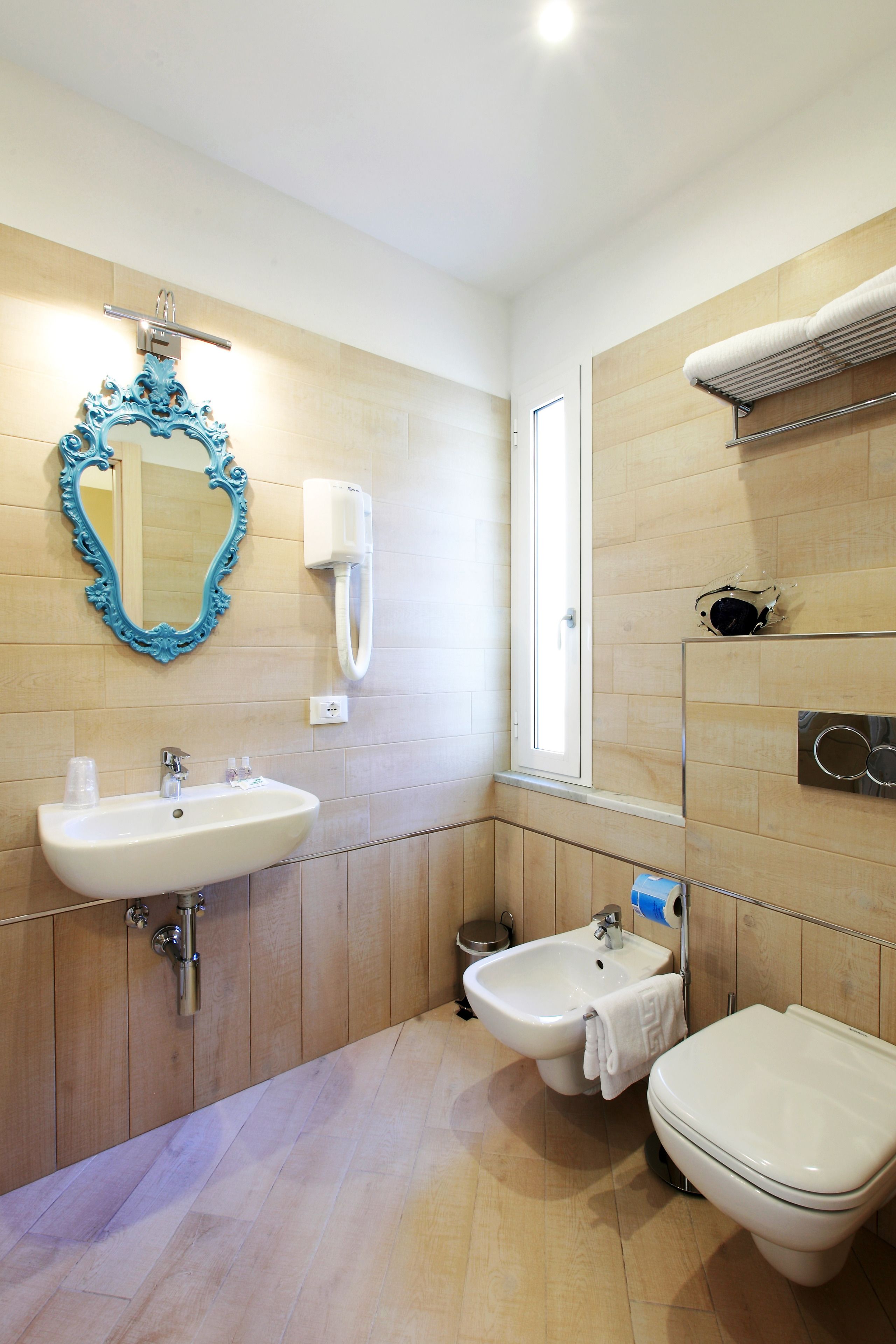 A bathroom with a sink, bidet, and toilet, wooden paneling, a mirror with an ornate frame, and a window.