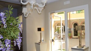 Vestíbulo de hotel con candelabro, una planta en maceta, lámpara y puerta de vidrio que da al exterior.