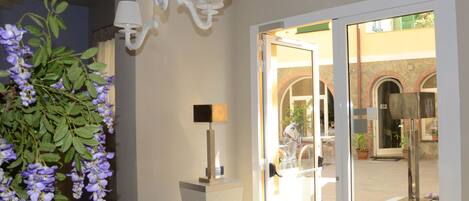 A hotel lobby with a chandelier, a potted plant, a lamp, and a glass door leading outside.