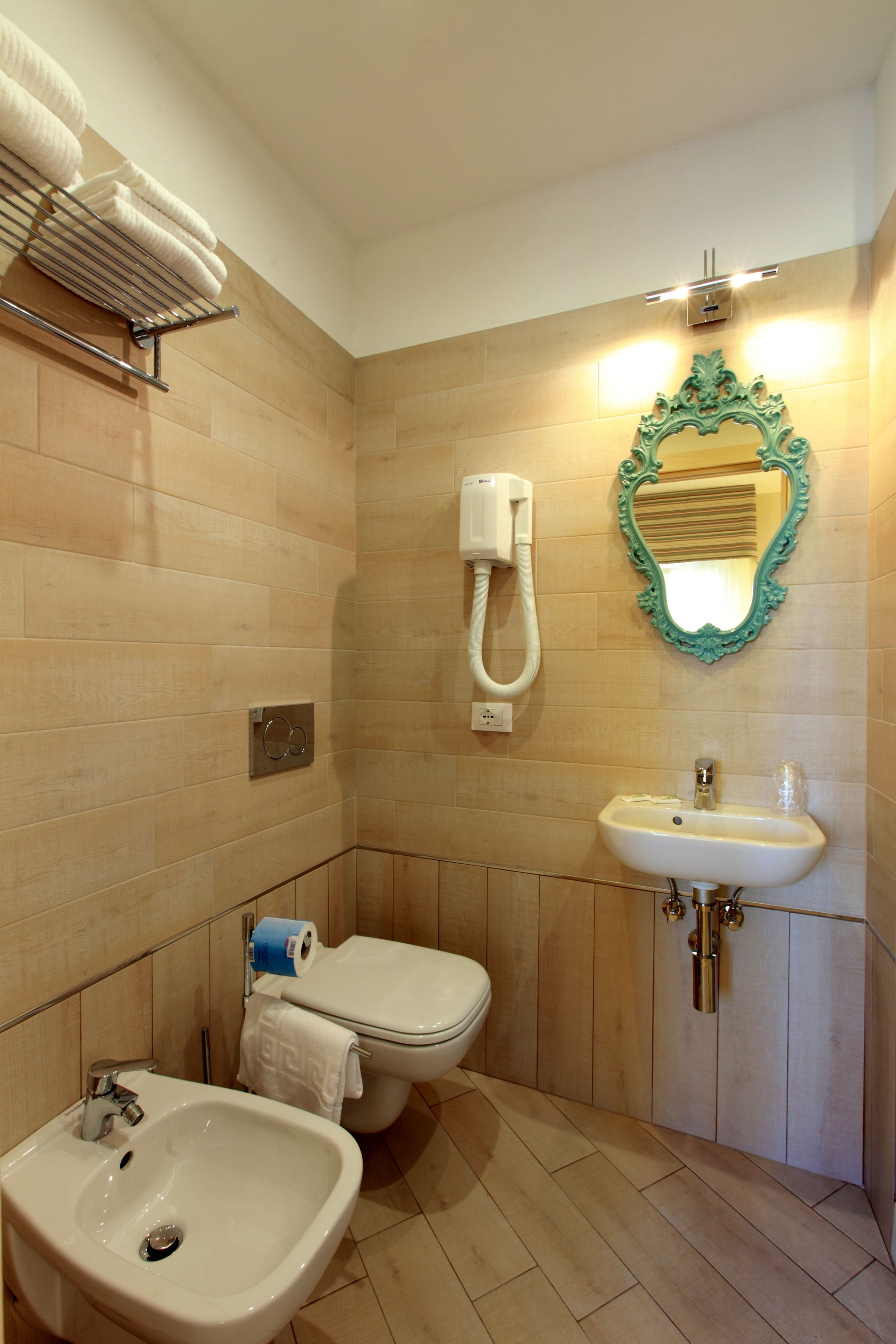 Une salle de bain équipée d’un lavabo, de toilettes et d’un bidet. On y trouve un porte-serviettes avec des serviettes, un miroir et un luminaire placé au-dessus du miroir.