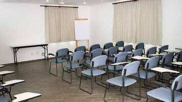 Uma sala de conferências com fileiras de cadeiras e mesas, um quadro branco e cortinas.