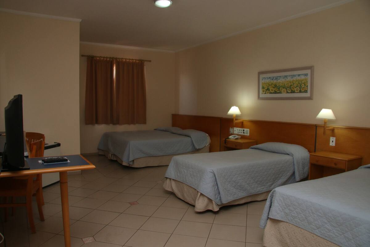 Habitación de hotel con dos camas individuales, un escritorio con computadora, televisor y ventana con cortinas.