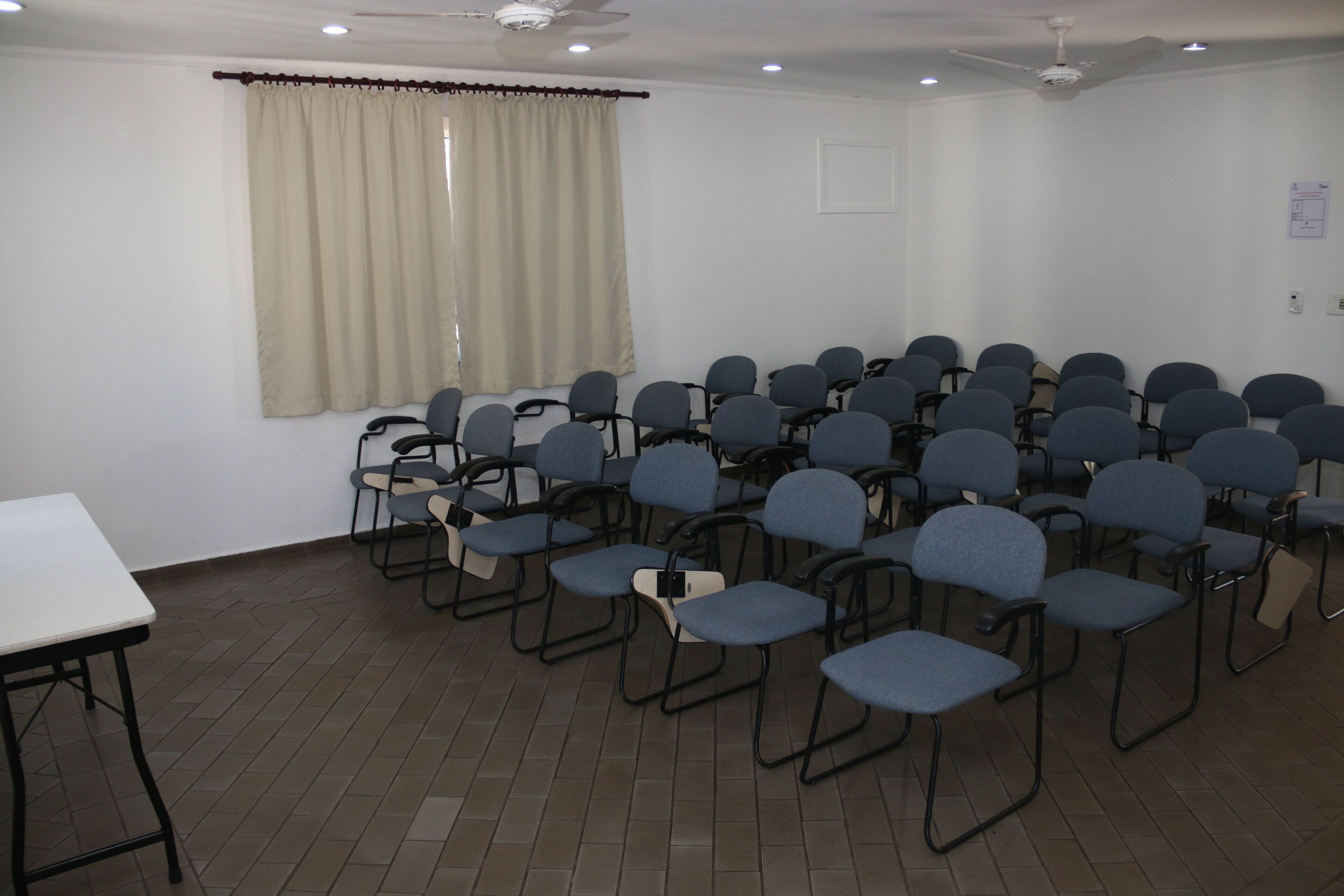 Uma sala de conferências com fileiras de cadeiras, uma mesa, um ventilador de teto e uma janela com cortinas.