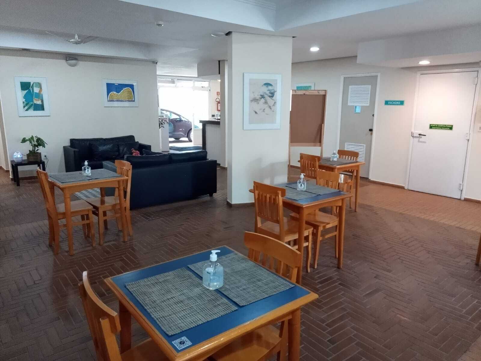 Un hall de hotel con una recepción, un sofá negro, mesas de madera con sillas y un piso de baldosas.