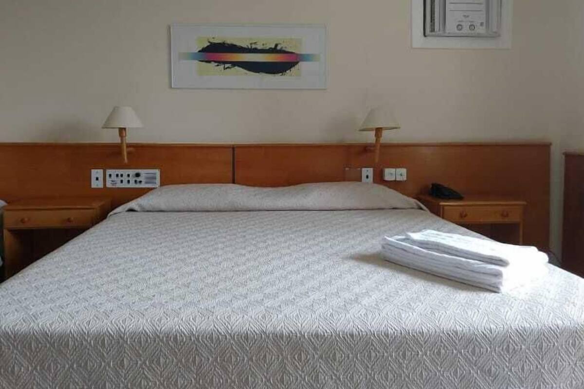 Habitación de hotel con una cama grande, dos mesitas de noche con lámparas, un equipo de aire acondicionado en la pared y un cuadro abstracto enmarcado.