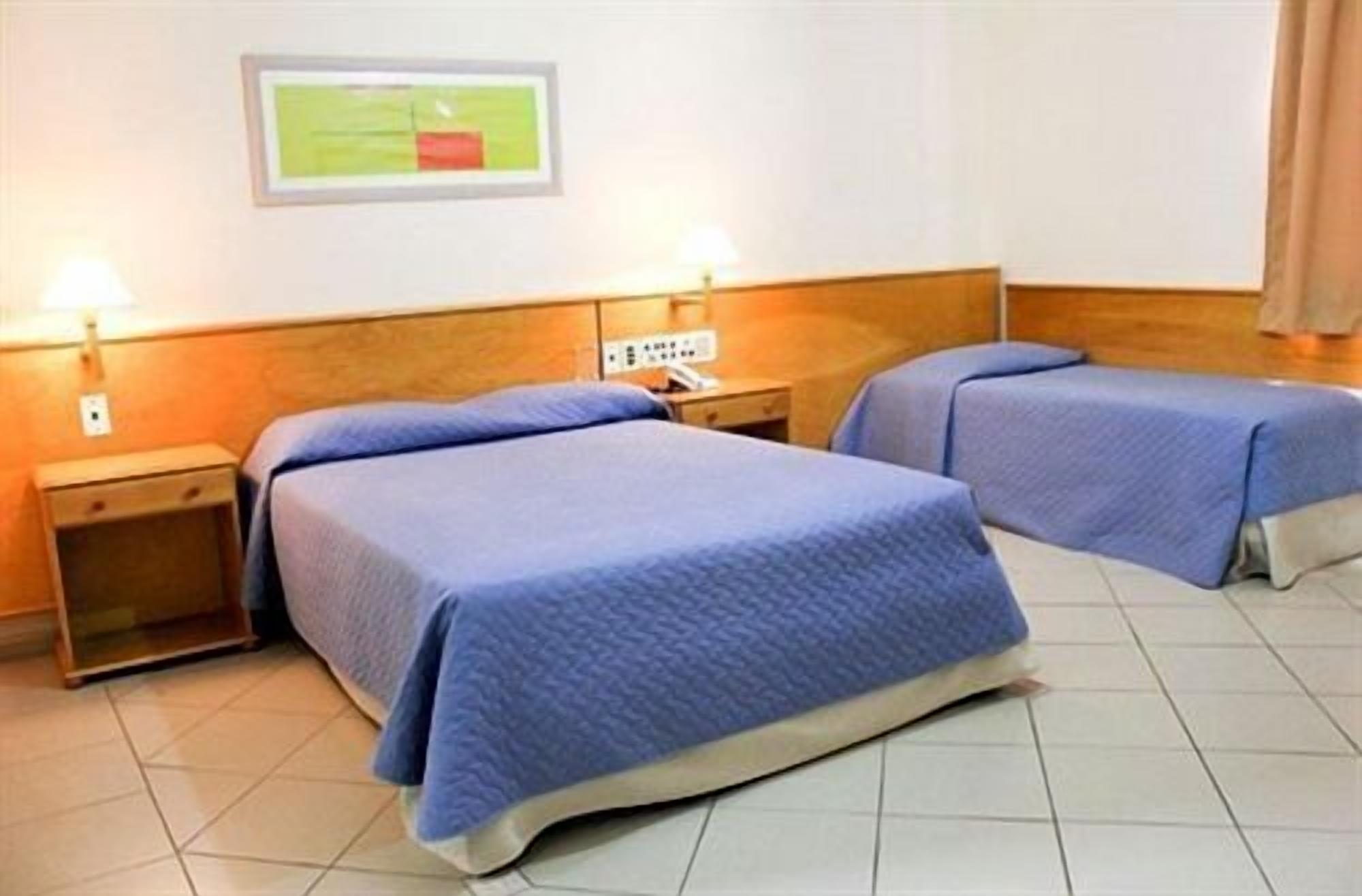 A hotel room with two beds, each with a wooden headboard and a blue bedspread. There is a small bedside table with a lamp on each side of the beds.